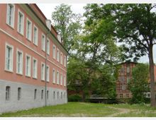 Großes historisches Gebäude mit rosa Fassade und weißen Fensterrahmen, umgeben von grünen Bäumen und Rasenflächen. Im Hintergrund ist ein Fachwerkhaus aus rotem Backstein zu sehen, das von einem kleinen Fluss oder Graben mit einer Brücke getrennt ist.