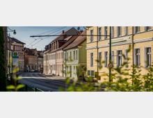 Straßenszene mit Reihenhäusern in Gelb- und Pastelltönen, teils mit klassischer Fassade und Satteldächern. Im Vordergrund sind eine Bushaltestelle, eine Geschwindigkeitsbegrenzung von 30 km/h und Oberleitungen für öffentliche Verkehrsmittel zu sehen.