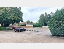 Ein leerer Parkplatz mit vereinzelten parkenden Autos, umgeben von Bäumen und Sträuchern. Im Hintergrund sind Ziegelmauern, ein überdachter Unterstand und Wohngebäude zu sehen.