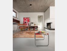 Modern minimalist living room with a raw concrete ceiling, white walls, and a white brick fireplace with a dark hearth. A low bookcase with framed art sits against the wall beside chrome-framed chairs on a rug, with a large window and doorway leading to another room.