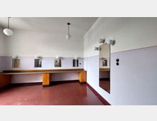 Ein schlicht eingerichteter Raum mit pastell-lila Wänden und einem roten Boden. An der Rückwand befindet sich ein langer Holztisch mit mehreren kleinen rechteckigen Spiegeln und Kugellampen darüber, an der Seitenwand ein großer rechteckiger Spiegel mit weiteren Wandlampen.