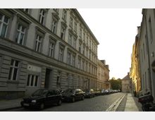 Pflastersteinstraße mit parallel geparkten Autos und mehrstöckigen Wohnhäusern im Altbaustil auf beiden Seiten. Sonnenlicht erleuchtet die rechte Gebäudeseite, während kleinere Bäume im Hintergrund sichtbar sind.