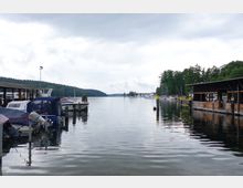 Hafenansicht mit Booten am Steg in einer ruhigen Gewässerbucht, umgeben von Waldufern. Rechts im Bild befinden sich Holzhütten mit überdachten Bereichen, und im Hintergrund sind weitere Boote und Segelboote sichtbar.