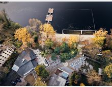 Aerial view of a lakeside area with a small dock extending into the water, surrounded by autumn trees. The scene includes a courtyard with scattered seating, landscaped paths, and several buildings with sloped roofs.