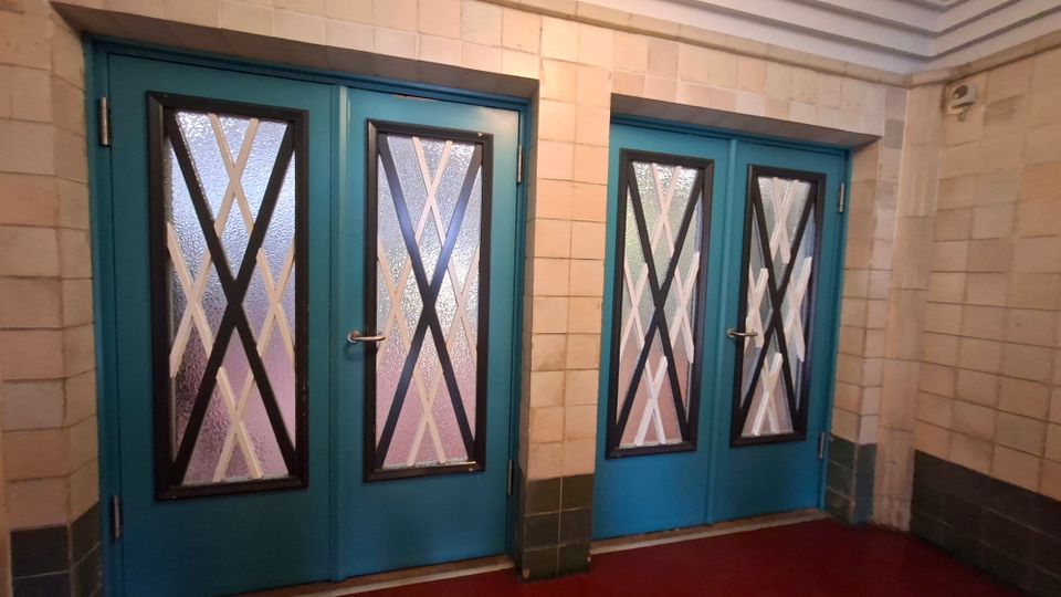 Eingangsbereich mit hellen, quadratischen Wandfliesen und zwei türkisfarbenen Doppeltüren mit schmalen Glasfenstern, die durch schwarze und weiße X-förmige Streben gegliedert sind. Roter Bodenbelag und dunklere Fliesen als Sockelzone entlang der Wände.