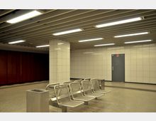Unterirdischer U-Bahnbereich mit weißen gefliesten Wänden, einer Metalltür, einer Reihe von Metallbänken und einem Mülleimer im Vordergrund. Die abgehängte Decke ist mit langen Leuchtstofflampen ausgestattet.