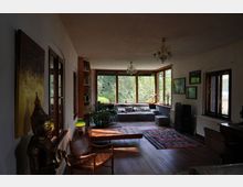 Helles Wohnzimmer mit großen Holzrahmenfenstern, die einen Blick auf einen grünen Garten bieten. Der Raum ist mit einem grauen Ecksofa, Holzmöbeln, Teppichen und dekorativen Elementen wie einer goldenen Buddha-Statue ausgestattet.