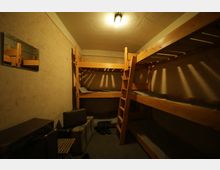 Kleiner Raum mit vier Holz-Etagenbetten, beleuchtet von einer Deckenlampe. An der linken Wand hängt ein Spiegel, daneben befinden sich Koffer und Schuhe auf dem Boden.