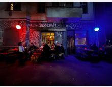 Straßenansicht eines Eingangs zu einer Bar namens „BERNDHAIN“ mit beschmierter Fassade und Graffiti, beleuchtet von roten und blauen Außenlampen. Vor dem Eingang stehen mehrere Holztische mit Bänken auf dem Gehweg, dahinter sind Fenster und ein kleiner Balkon an der Gebäudefront zu sehen.