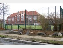Backsteingebäude mit weißen Verzierungen und rotem Satteldach, vermutlich eine Schule, im Hintergrund. Im Vordergrund ein umzäunter Sportplatz mit grünem Kunstrasen, umgeben von kahlen Sträuchern und vereinzelten Baumstämmen.