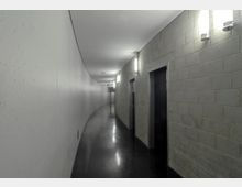 Ein langer, schmaler Flur mit einer geschwungenen Wand, einer Seite aus glatten, weißen Flächen und einer gegenüberliegenden Seite aus grauen Ziegelmauern mit Türen. Der Boden ist schwarz glänzend, und die Beleuchtung erfolgt durch Wandleuchten an der Ziegelwand.