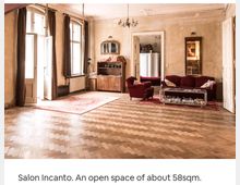 Ein historischer Salon mit offenem Raum, Parkettboden und hohen Decken. Der Raum ist mit roten Polstermöbeln, Teppichen, einem Holzschrank und großen Fenstern ausgestattet, die viel Tageslicht einlassen.