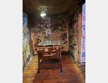 Kleiner, schmaler Innenraum mit Holzboden und niedriger Decke, deren Wände dicht mit Graffiti, Stickern und Kritzeleien bedeckt sind. In der Mitte steht ein Kickertisch (Original Leonhart) unter einer Deckenlampe.