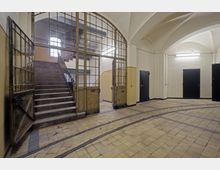 Ein historisches Treppenhaus mit hohen Decken und Rundbögen. Sichtbar sind eine breite Steintreppe hinter einer verglasten Metalltür sowie ein Fliesenboden mit eingelassenen Schienen; im Hintergrund mehrere dunkle Türen.