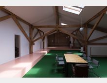 Großer, moderner Dachraum mit sichtbaren Holzbalken und Oberlichtern, ausgestattet mit einem langen Holztisch, umgeben von Stühlen auf einem grünen Teppichboden. Wände sind teilweise weiß, teilweise mit einer Backsteinwand, und der Boden enthält glänzend rosa Bereiche.