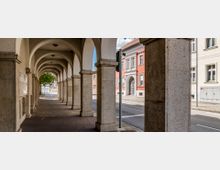 Arkaden eines Gebäudes mit einem überdachten Gehweg, gehalten von massiven Steinpfeilern und Rundbögen. Im Hintergrund ist eine Straße mit historischen Fassaden, darunter ein rotes Gebäude, zu sehen.