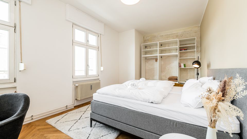 Helles Schlafzimmer mit großem grauem Polsterbett, weißen Wänden und hohen Fenstern mit Heizkörper darunter. Im Hintergrund stehen offene Regale vor einer roh verputzten Wand; Holzboden im Fischgrätmuster, kleiner Teppich und Nachttischlampe ergänzen die Einrichtung.