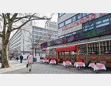 Straßenszene mit einem italienischen Restaurant namens "Capone" an einem Gebäude mit der Aufschrift "Hollywood Media". Vor dem Restaurant stehen Tische mit rot-weiß karierten Tischdecken und Stühlen, auf einem Gehweg mit vorbeigehenden Passanten und kahlen Bäumen.