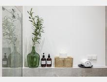 Modernes Badezimmerdetail mit Glasduschwand und grauen Fliesen; auf einer Ablage stehen eine große grüne Glasvase mit Zweig, zwei Spenderflaschen, ein geflochtener Korb mit Toilettenpapier und ein aufgerolltes Handtuch auf einer Schale vor weißer Wand mit Lichtschaltern.