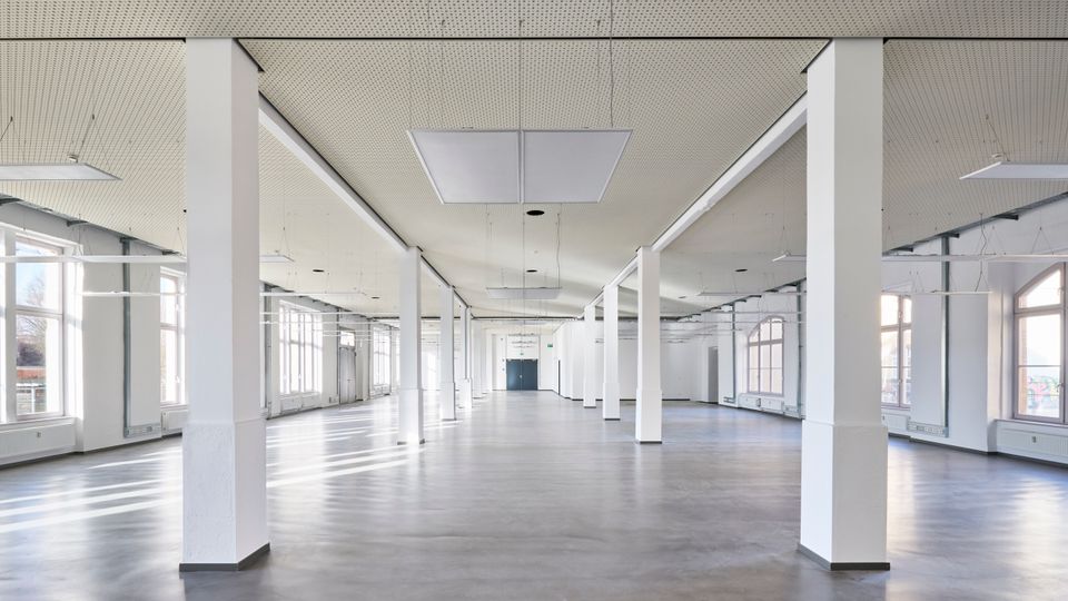 Großer, leerer Innenraum mit glattem Betonboden, weiß gestrichenen Wänden und mehreren quadratischen Säulen. Die rechteckigen Fenster entlang der Wand lassen viel Tageslicht herein, während die Decke mit eingelassenen Lichtfeldern und perforierten Paneelen gestaltet ist.