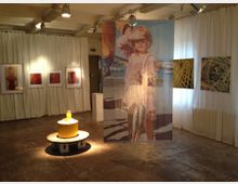 Galerieraum mit hellen weißen Vorhängen an den Wänden, die mit Fotografien und abstrakten Kunstwerken geschmückt sind. Im Vordergrund steht eine gelbe skulpturale Installation auf einem runden Podest, während ein großformatiges Foto eines Kindes im Zentrum hängt.