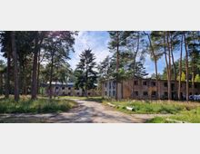 Zwei verlassene, zweistöckige Gebäude mit beschädigten Fassaden stehen inmitten eines lichten Kiefernwaldes. Ein schmaler Weg führt zwischen hohen Bäumen und Wiesenflächen zu den Gebäuden, während Wäschestücke an einer Leine rechts im Bild hängen.