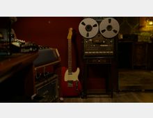 Aufnahme eines Musikstudios mit einer roten E-Gitarre, die an einer dunklen Wand lehnt, und einem großen Bandgerät mit zwei Spulen auf einem verzierten Tisch. Im Vordergrund sind Mischpulte und Audioequipment sichtbar, während der Raum eine warme, gedämmte Beleuchtung zeigt.