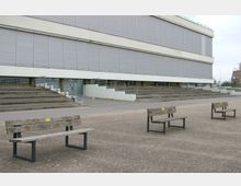 Moderne Gebäudefassade mit grauen Fensterläden, umgeben von einer gepflasterten Fläche. Im Vordergrund stehen mehrere einfache Holzbänke mit Metallgestell, seitlich führen Treppenstufen zum Eingang des Gebäudes.