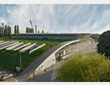 Modernes, langgestrecktes Gebäude mit geschwungener Glasfassade und begrüntem Dach, umgeben von Bäumen; im Hintergrund stehen hohe Lichtmasten eines Stadions.