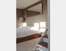 Minimalistisch eingerichtetes Schlafzimmer mit einem niedrigen Holzbett und weißer Bettdecke, einem rechteckigen Spiegel an einer beigefarbenen Wand und einer einfachen, hölzernen Treppe im Vordergrund. Natürliches Licht fällt durch ein Fenster ins Zimmer.