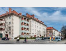 Eine Straßenansicht mit mehrstöckigen Wohngebäuden aus der Nachkriegszeit, die helle Fassaden und rote Ziegeldächer aufweisen. Im Vordergrund fahren Radfahrer, während Autos am Straßenrand geparkt sind; vereinzelte Bäume säumen die Straße.