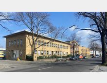 Backstein-Gebäude mit zwei Etagen entlang einer Straße, mit zahlreichen Fenstern und einem Flachdach; im Vordergrund stehen Autos und ein Anhänger geparkt, während kahle Bäume die Szenerie säumen. Der Himmel ist blau mit einigen Wolken.