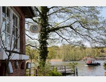 Backsteingebäude mit weißen Sprossenfenstern am Ufer eines Sees, umgeben von Bäumen und Sträuchern. Im Hintergrund eine kleine Fähre auf dem Wasser sowie ein Holzsteg am Ufer.