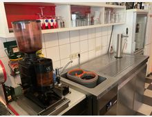 Getränkezubereitungs- und Spülbereich mit Edelstahlarbeitsplatte, integrierter Spüle und Abtropffläche sowie Zapfhahn; daneben steht eine Kaffeemühle. Darüber befinden sich offene Regale mit Gläsern und Tassen vor einer weiß gefliesten Wand.