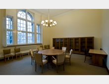 Ein Besprechungsraum mit einer hohen Decke und großen Fenstern, die durch Holzrahmen unterteilt sind. Ein runder Holztisch mit Stühlen steht im Zentrum, an den Wänden befinden sich Holzvitrinen und zusätzliche Stühle.