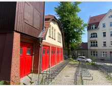 Roter Klinkerbau mit einem hell verputzten Obergeschoss und mehreren roten Toren, die an eine historische Feuerwache erinnern. Im Vordergrund sind Fahrradständer auf einem gepflasterten Platz, dahinter Bäume und benachbarte Wohngebäude.