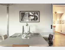 Ein modernes Büro mit einem großen grauen Tisch, darauf Monitore, eine Tischlampe und ein Telefon. An der Wand hängt ein gerahmtes schwarz-weißes Kunstwerk; im Hintergrund ist ein Gang mit gerahmten Bildern an den Wänden sichtbar.