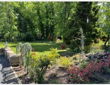 Ein Garten mit einer Rasenfläche, umgeben von dichten Bäumen und Sträuchern. Im Vordergrund sind ein gepflasterter Weg, eine kleine Steinmauer, eine Laterne sowie bunte Pflanzen und Blumenbeete sichtbar.