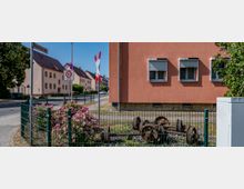 Ecke einer Wohnstraße mit einem lachsfarbenen Gebäude, geschlossenen Rollläden und einem kleinen bepflanzten Bereich vor einem Zaun, in dem alte Eisenbahnräder liegen. Im Hintergrund sind weitere Wohnhäuser und Verkehrszeichen zu sehen.