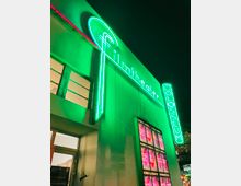 Fassade eines Kinos bei Nacht mit grün leuchtenden Neonbuchstaben "Filmtheater Colosseum" und einem leuchtenden Kinoprogramm an der Wand. Ein Fenster und ein Eingang sind ebenfalls zu erkennen.