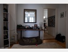 Ein Flur mit hellem Holzboden und weiß gestrichenen Wänden. Im Vordergrund ein Hundebett auf einem Teppich, darüber ein schmaler Holz-Tisch mit persönlichen Gegenständen und einer Lampe, sowie ein an der Wand hängender Spiegel, der ein Fenster reflektiert. Rechts führt eine Tür in einen weiteren Raum mit Holzmöbeln und einer Pinnwand. Links steht ein Regal mit Schuhen.