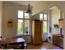 Helle Küche mit Holzmöbeln, einem Esstisch mit Stühlen, einer Waschmaschine und einer mit gemusterten Fliesen verkleideten Arbeitsfläche. Große Fenster lassen Tageslicht herein und bieten Blick auf grüne Bäume.