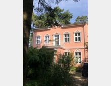 Ein zweistöckiges Haus in Altbauarchitektur mit rosa Fassade, weißen Fensterrahmen und einem Balkon im Obergeschoss. Umgeben von dichter Vegetation, darunter Bambus und blühende Büsche; im Vordergrund steht ein geparktes Auto.
