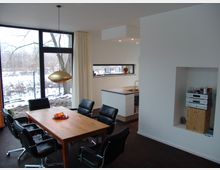 Moderner Wohnraum mit Esstisch und Küche. Ein Holztisch mit schwarzen Stühlen steht vor einer großen Fensterfront mit Blick auf verschneites Außengelände, daneben eine kleine Küche mit weißer Einrichtung und länglichem Fenster.