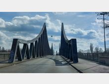 Eine moderne Brücke mit geschwungenen Stahlbögen und diagonalen Streben, die über eine Straße gespannt ist. Im Hintergrund sind Bäume und ein teils bewölkter Himmel zu sehen.