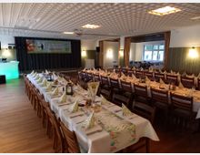 Ein festlich dekorierter Veranstaltungsraum mit langen, gedeckten Tischen und Holzstühlen. Im Hintergrund befinden sich eine Bühne mit einem Banner sowie grün gestrichene Wände und Fenster, die Tageslicht hereinlassen.