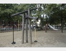 Spielplatz mit Holzspielgeräten, darunter eine Konstruktion mit Schaukeln und Seilen, sowie eine Rutsche und ein Kletternetz im Hintergrund; umgeben von Bäumen und einer umzäunten Umgebung. Im Hintergrund sind Wohnhäuser erkennbar.