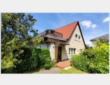 Einfamilienhaus mit Satteldach und roter Dacheindeckung, umgeben von dichtem Grün und Büschen. Das Haus hat eine kleine Veranda und einen Balkon im Obergeschoss.
