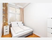 Helles, minimalistisches Schlafzimmer mit Doppelbett und weißer Bettwäsche, neben einem kleinen Nachttisch. Große Fenster mit beigefarbenem Vorhang und eine freiliegende Ziegelwand setzen Akzente, dazu Holzfußboden und weiße Wände.