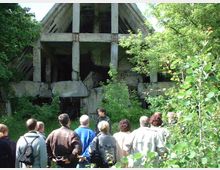 Eine Gruppe von Menschen steht vor einer verfallenen, überwucherten Betonstruktur mit offenen, zerstörten Wänden und sichtbaren Stahlträgern, umgeben von dichtem Grün aus Büschen und Bäumen.